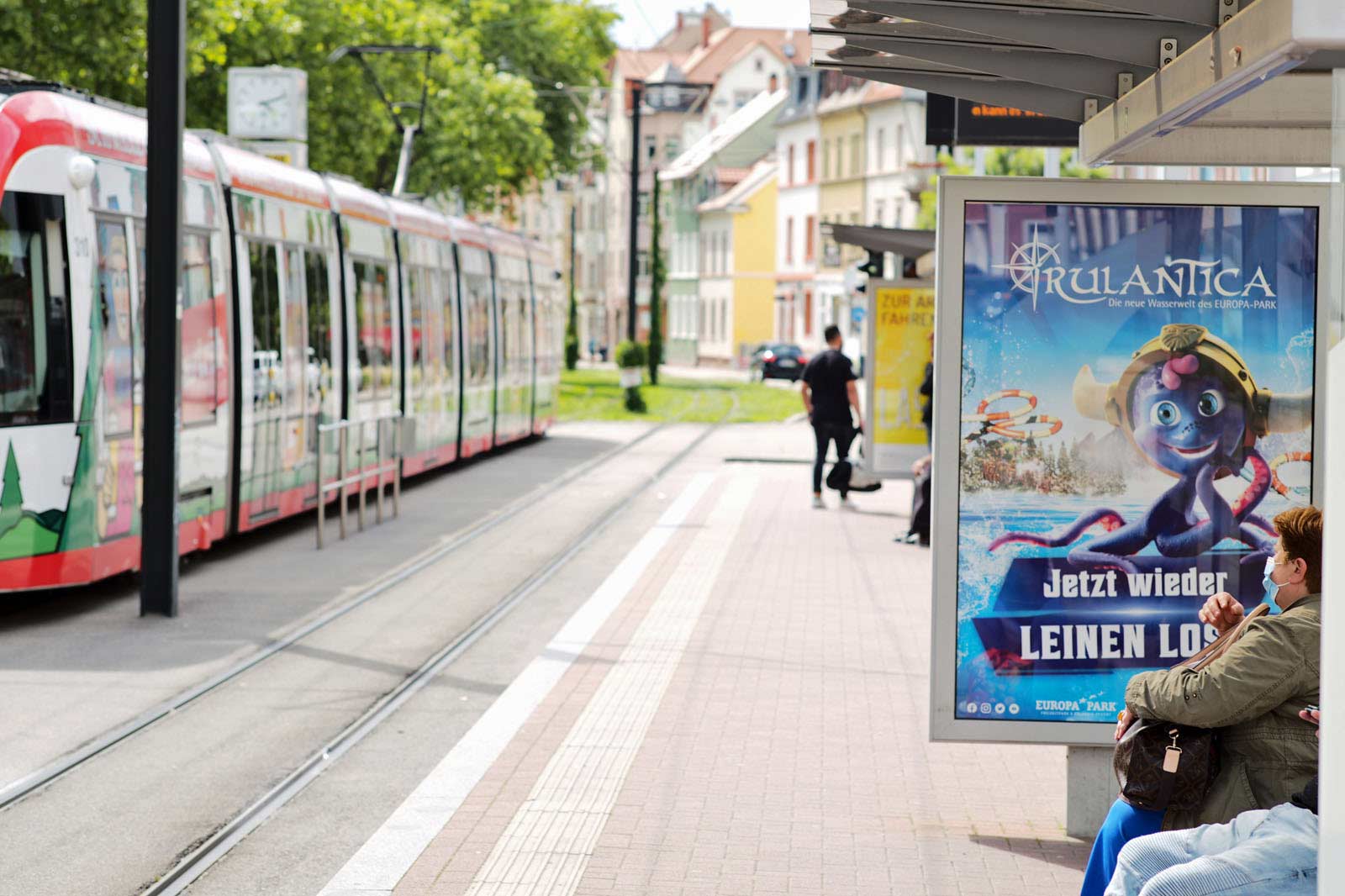 CLP in Fahrgastunterstand mit dem Motiv Rulantica, dabei passiert eine Straßenbahn mit Werbung der Brauerei Rothaus